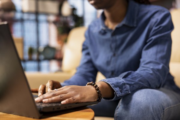 closeup of a person sitting on a couch leaning forward and typing on their laptop that is on the coffee table 